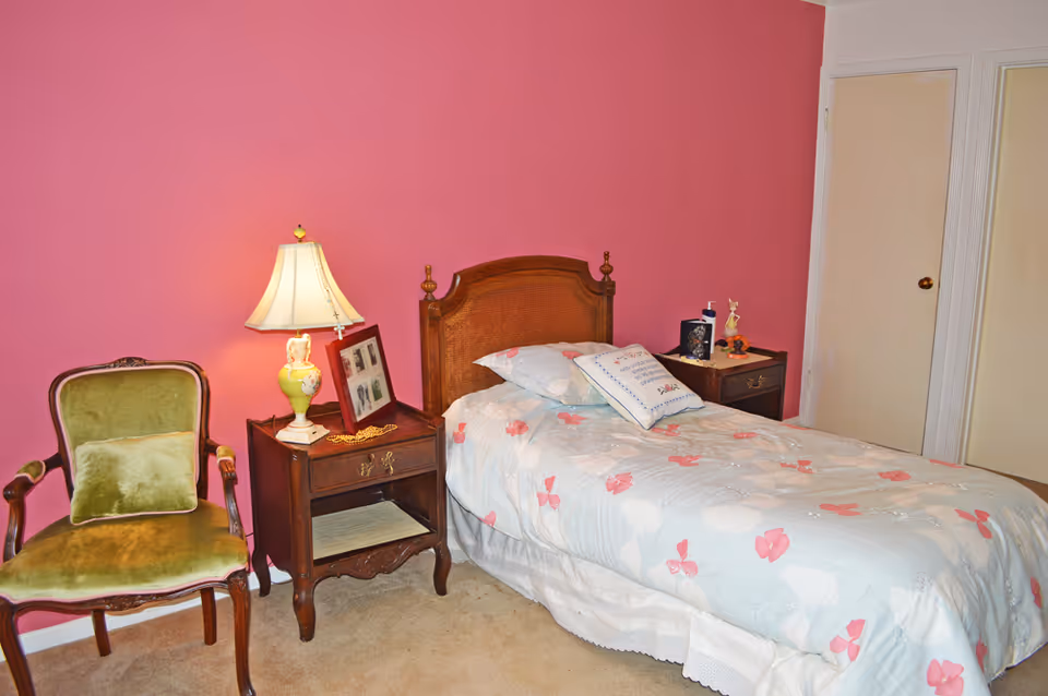 A bedroom with a single bed covered in a light blue and pink floral comforter, a wooden headboard, two wooden nightstands with decorative items and a lamp, a green upholstered chair with a matching pillow, and a pink accent wall behind the bed.