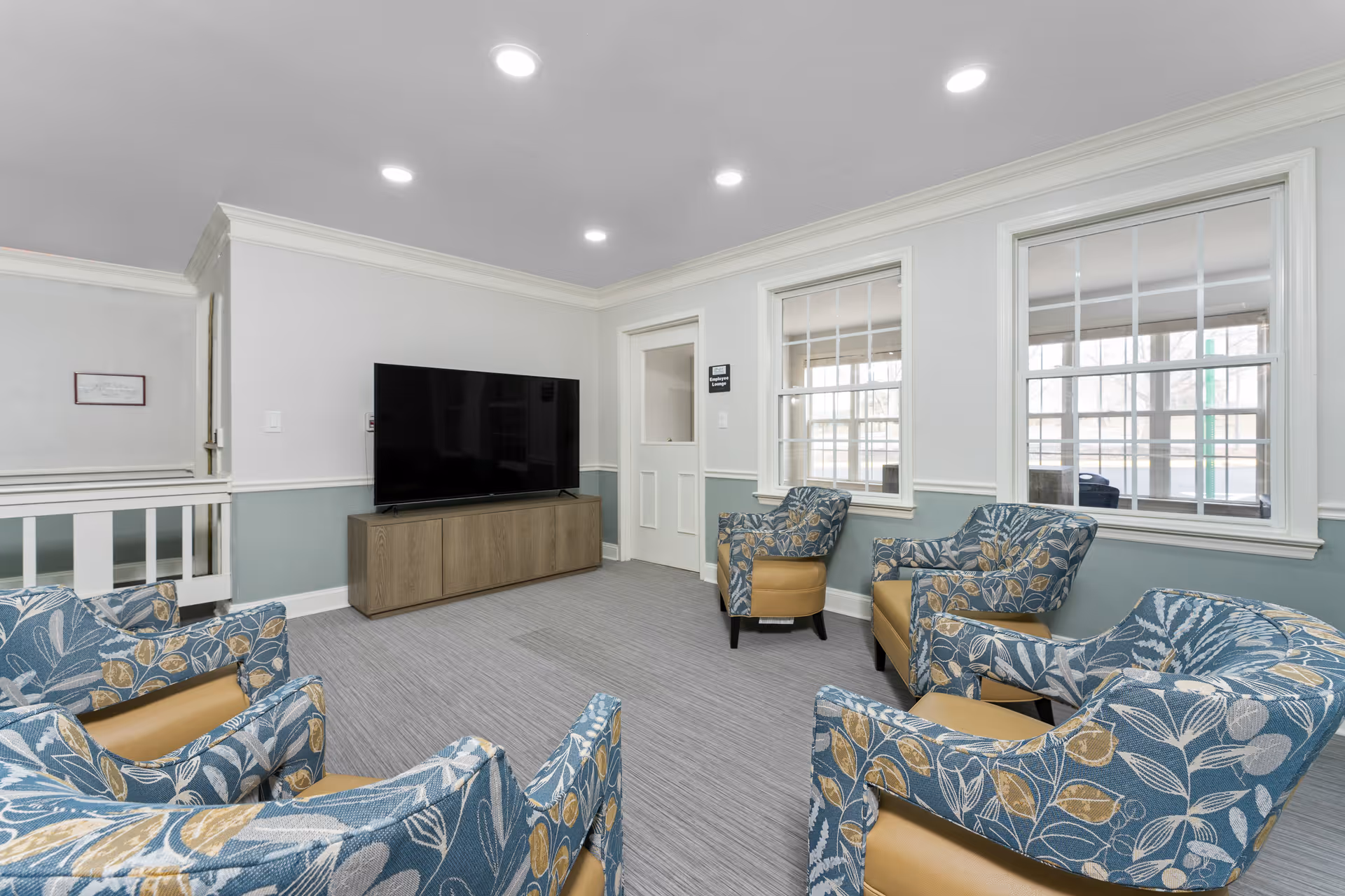 Bright common sitting room with patterned armchairs arranged facing a flat-screen TV on a wooden console and large windows.