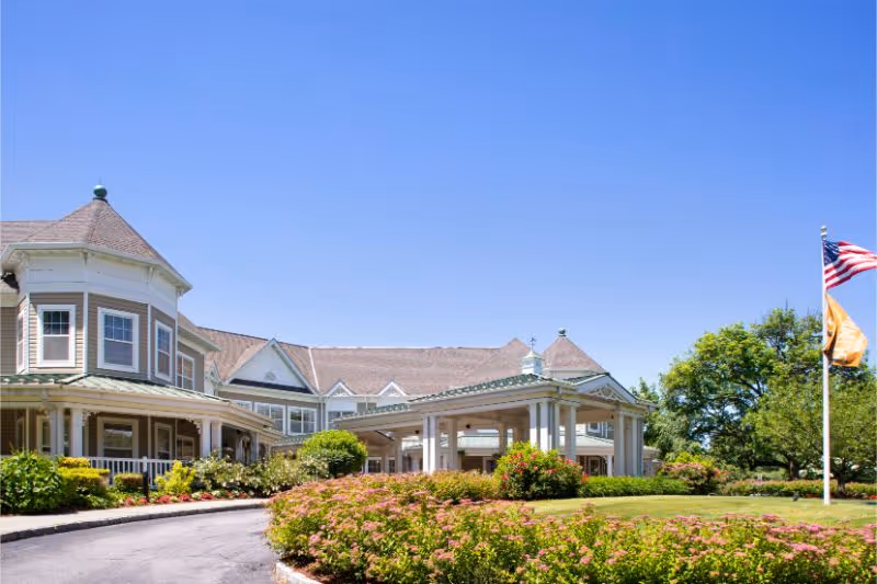 Front exterior of a senior living building with a covered entrance, circular driveway, landscaped flower beds, and flags under a clear blue sky.