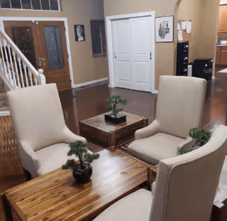 Interior view of a living room area with four beige upholstered chairs arranged around two wooden coffee tables. Small potted plants are placed on the tables. In the background, there is a staircase, a wooden front door with glass panels, a white double-door closet, and part of a kitchen area visible.