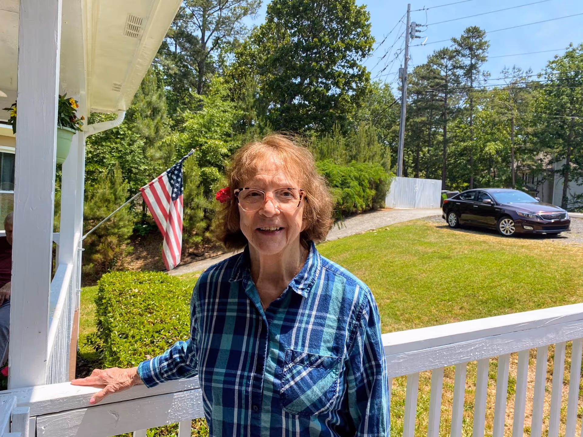 An elderly woman with glasses and a blue plaid shirt stands on a white wooden porch railing outside a building. Behind her is a green lawn, a black car parked on a gravel driveway, an American flag on a pole, and trees under a clear blue sky.
