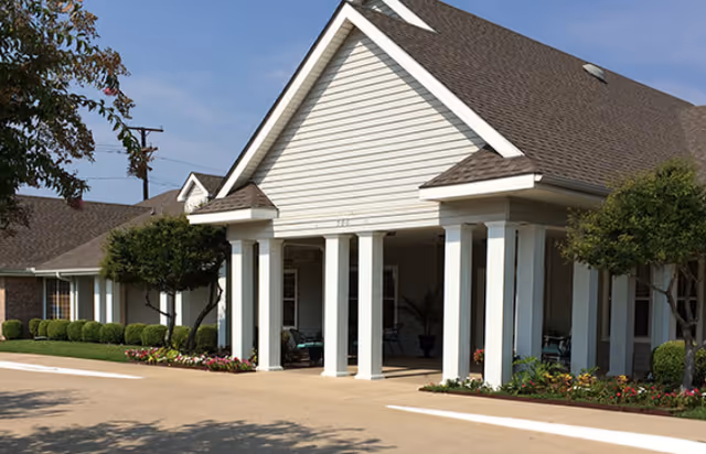 Front entrance of a single-story building with white columns, a covered porch, and landscaped flower beds.