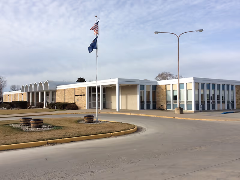 Exterior view of a single-story building with a flat roof and large windows. The building has a beige brick facade and a covered entrance supported by white columns. Two flags, one American and one state flag, are flying on a flagpole in front of the building. There is a curved driveway and a streetlamp near the entrance, with some landscaping including grass and wooden planters.