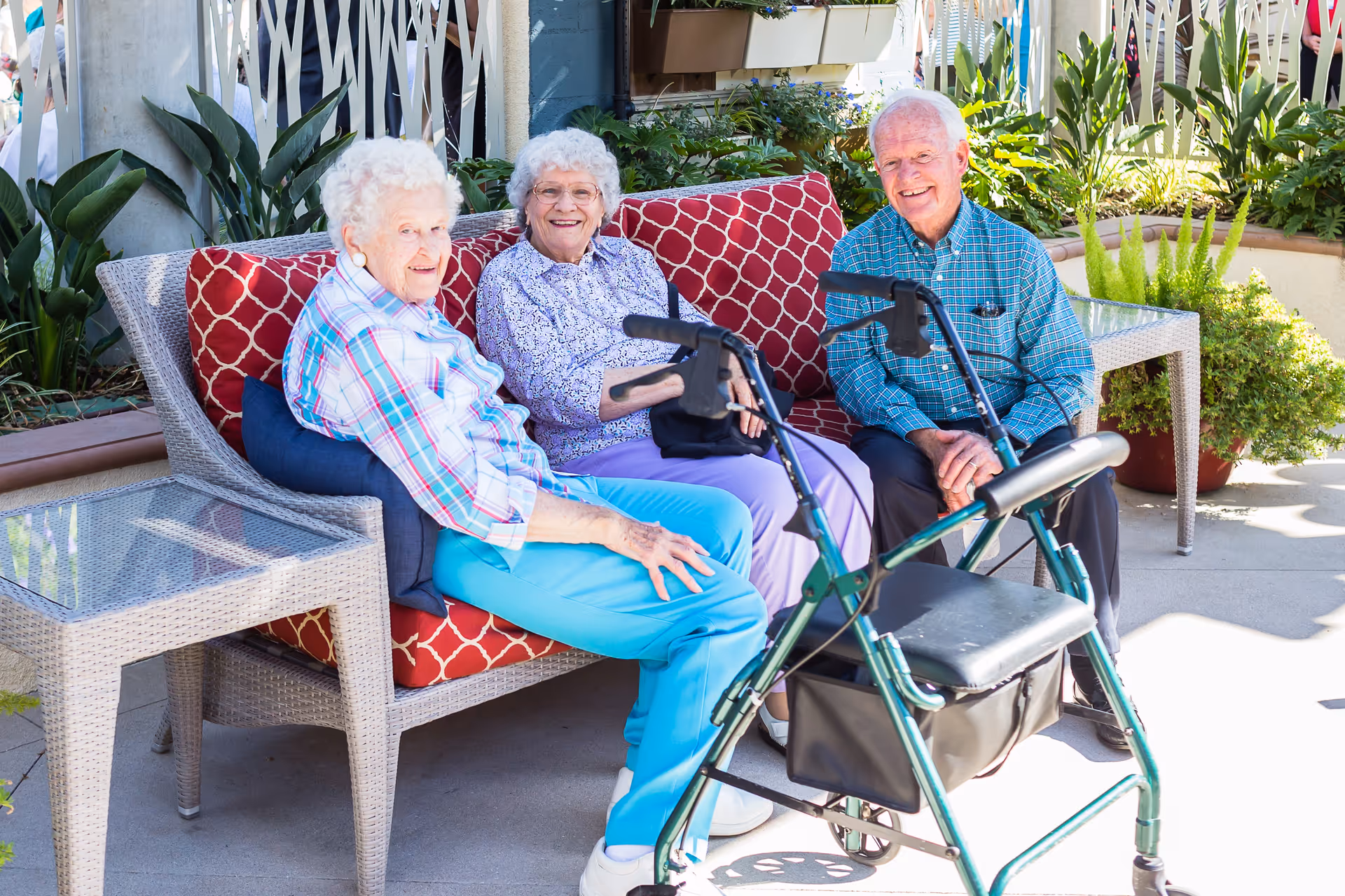 Three elderly individuals sitting on a cushioned outdoor wicker sofa with red patterned pillows, smiling and enjoying a sunny day. A green walker is positioned in front of them, and there are plants and a glass-top side table nearby.