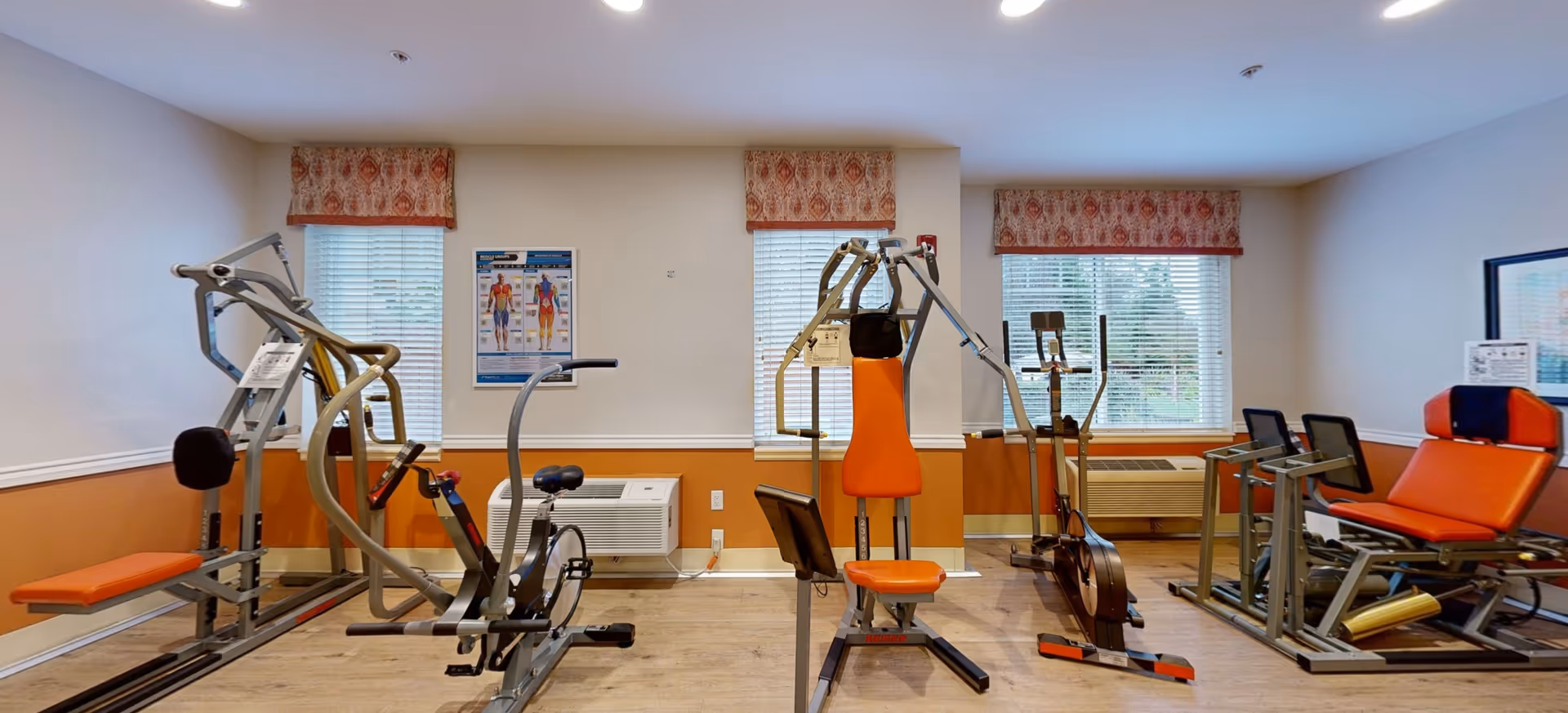 A fitness room with various exercise machines including a rowing machine, stationary bike, and weight training equipment. The room has three windows with patterned valances, light-colored walls with a chair rail, and wood flooring.