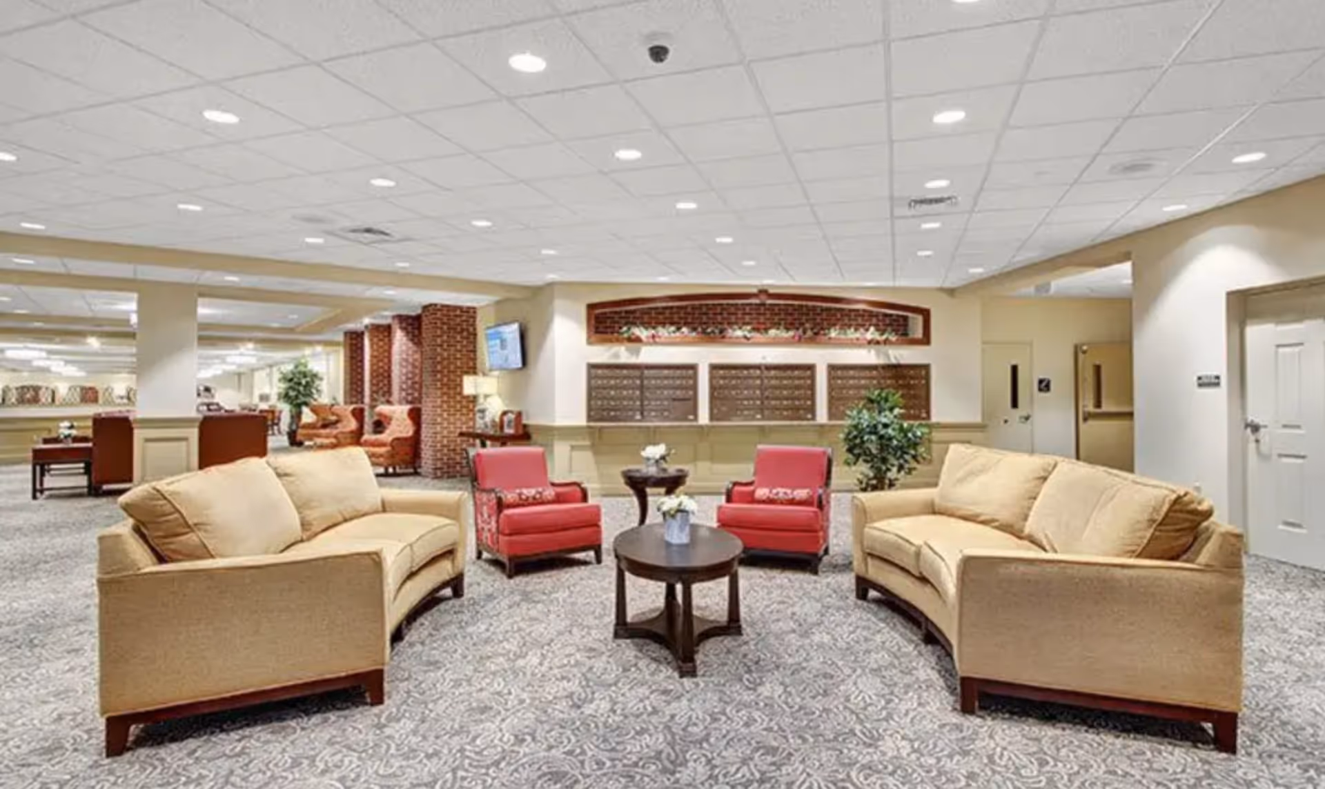 A spacious and well-lit common area with two beige sofas facing each other and two red armchairs with patterned cushions in between. A round wooden coffee table with a small flower arrangement is centered among the seating. The background features a wall with multiple mailboxes and decorative plants, along with a TV mounted on a brick column. The carpet has a subtle floral pattern, and the ceiling is fitted with recessed lighting.