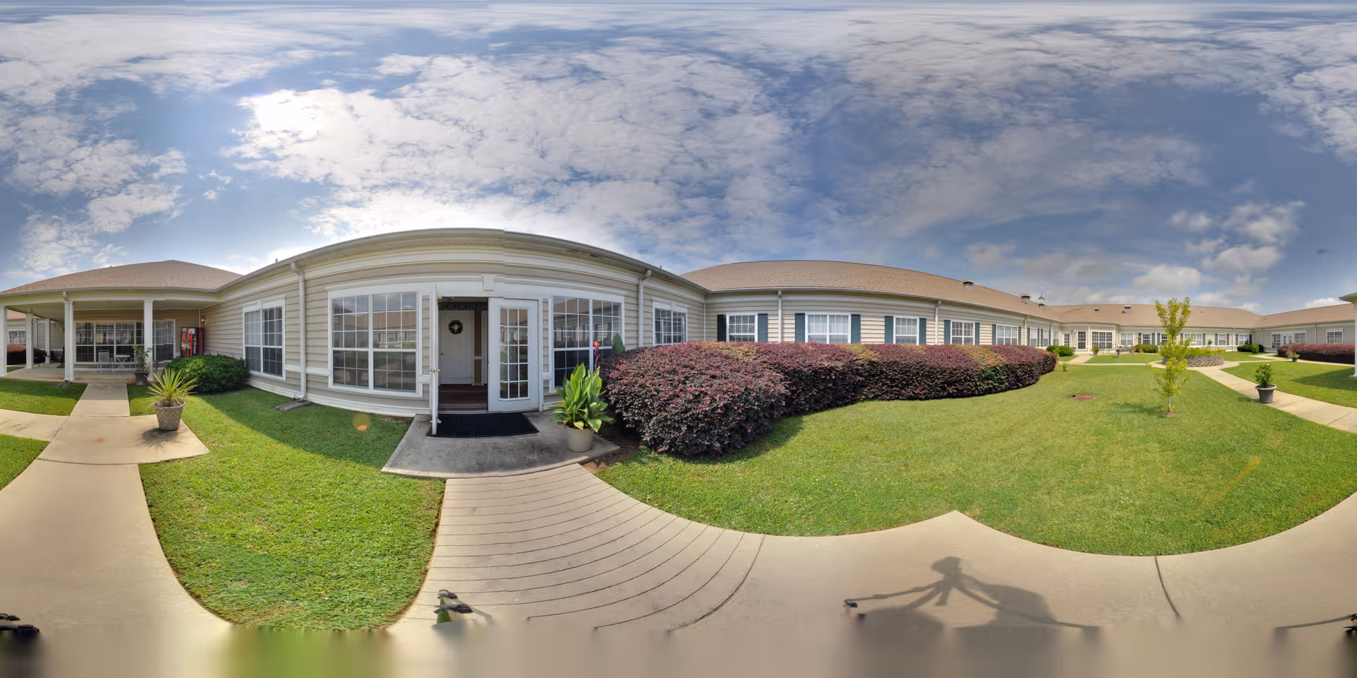 Wide panoramic view of the exterior courtyard of Oaks at Evans senior living facility, showing a single-story building with large windows, a covered entrance, well-maintained green lawns, shrubs, and a partly cloudy sky.