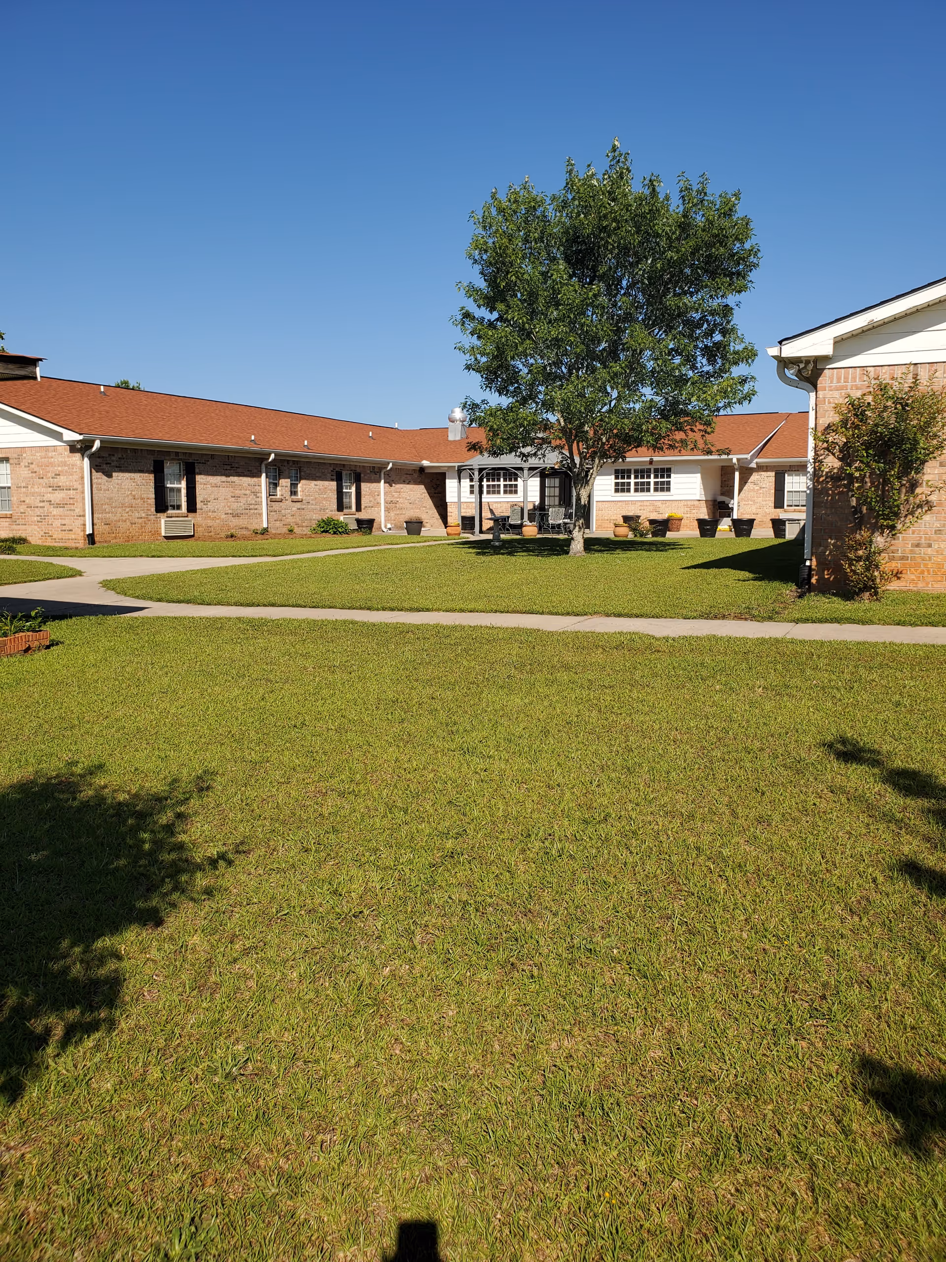A grassy courtyard area with a large tree in the center, surrounded by single-story brick buildings with red roofs under a clear blue sky.