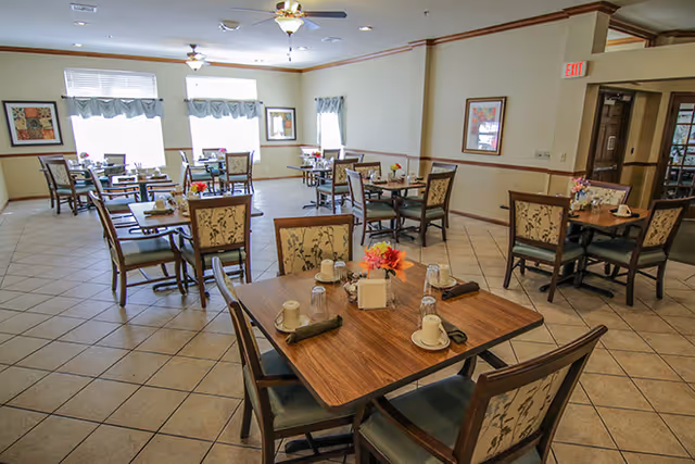 A bright dining room with multiple wooden tables and chairs arranged neatly. Each table is set with cups, glasses, napkins, and small floral centerpieces. The room has tiled floors, large windows with valances allowing natural light, and framed artwork on the walls.