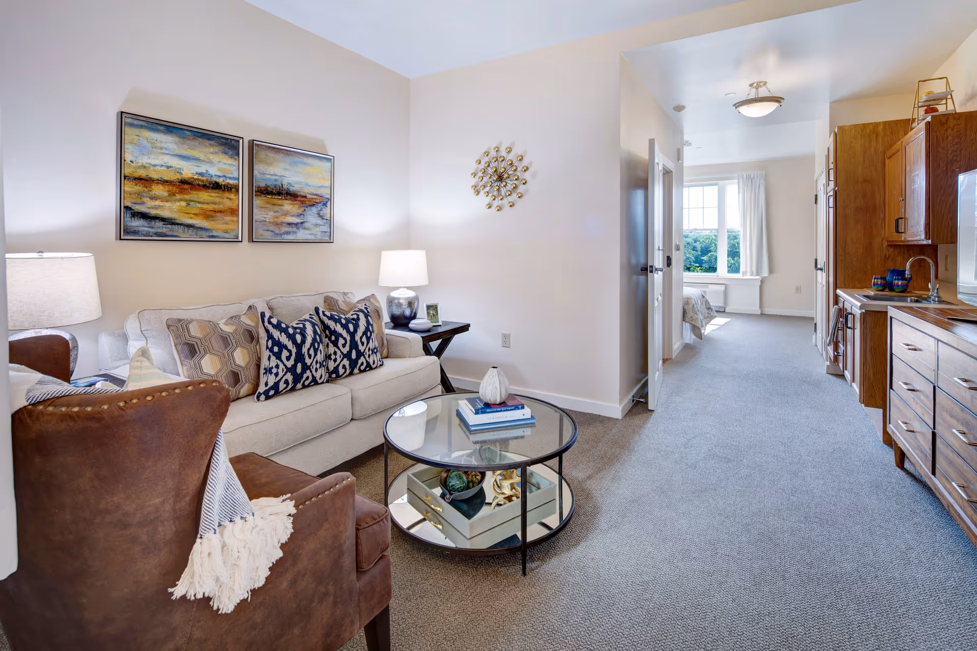 A cozy living area with a beige sofa adorned with patterned pillows, a brown armchair with a throw blanket, and a round glass coffee table with books and decorative items. The room has light-colored walls with two landscape paintings and a decorative wall clock. To the right, there is a kitchenette with wooden cabinets and a sink. In the background, a hallway leads to a bedroom with a large window letting in natural light.