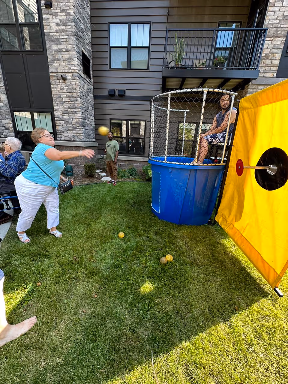 An outdoor scene at Amira Choice Bloomington showing a woman throwing a ball towards a dunk tank where a man is seated inside the tank. Other people are standing nearby watching the activity. The setting is a grassy area next to a building with stone and siding exterior walls and windows.