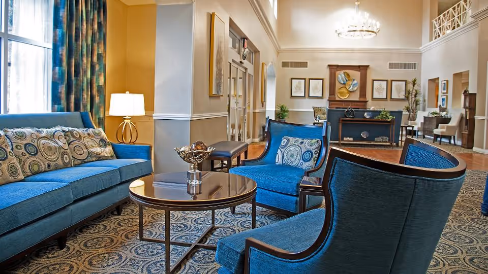A spacious and elegantly furnished senior living community lounge area with blue upholstered sofas and chairs arranged around a circular wooden coffee table. The room features patterned carpet, a decorative lamp, large windows with blue and green curtains, and a chandelier hanging from the ceiling. The background shows a fireplace with framed artwork and additional seating areas.