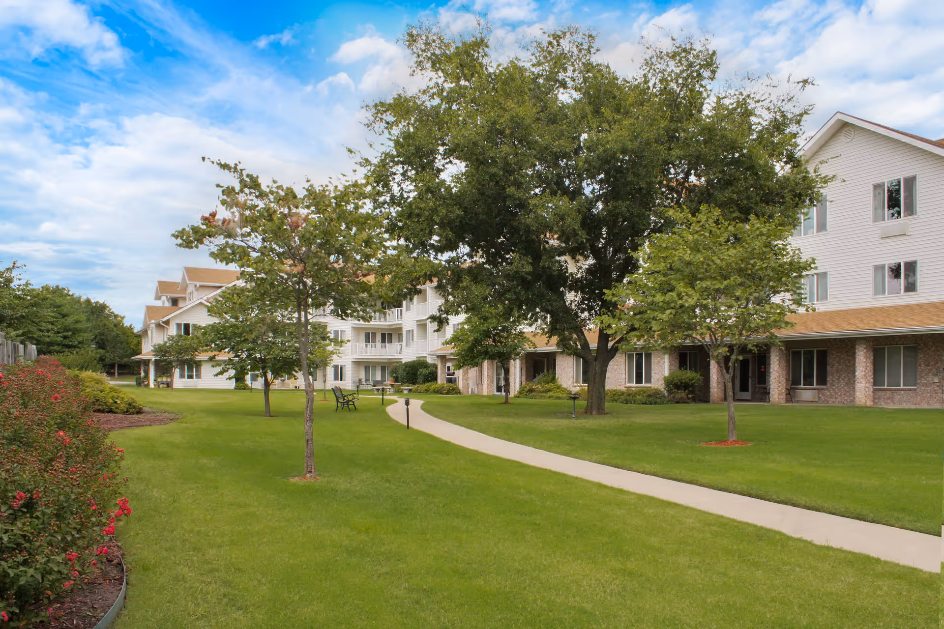 A well-maintained outdoor area of a senior living facility with a paved walkway, green grass, several trees, and benches. The multi-story building with white siding and brick accents is visible in the background under a partly cloudy sky.
