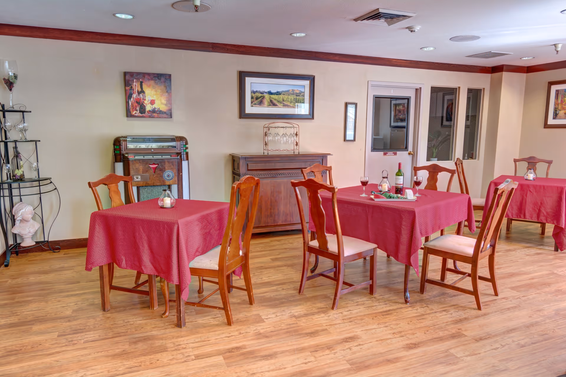 A dining room with wooden floors and several tables covered with red tablecloths. Each table is surrounded by wooden chairs with cushioned seats. On one table, there is a bottle of wine, two glasses of wine, and a small candle. The room has framed artwork on the walls, a vintage jukebox, and a decorative bust on a metal stand in the corner.