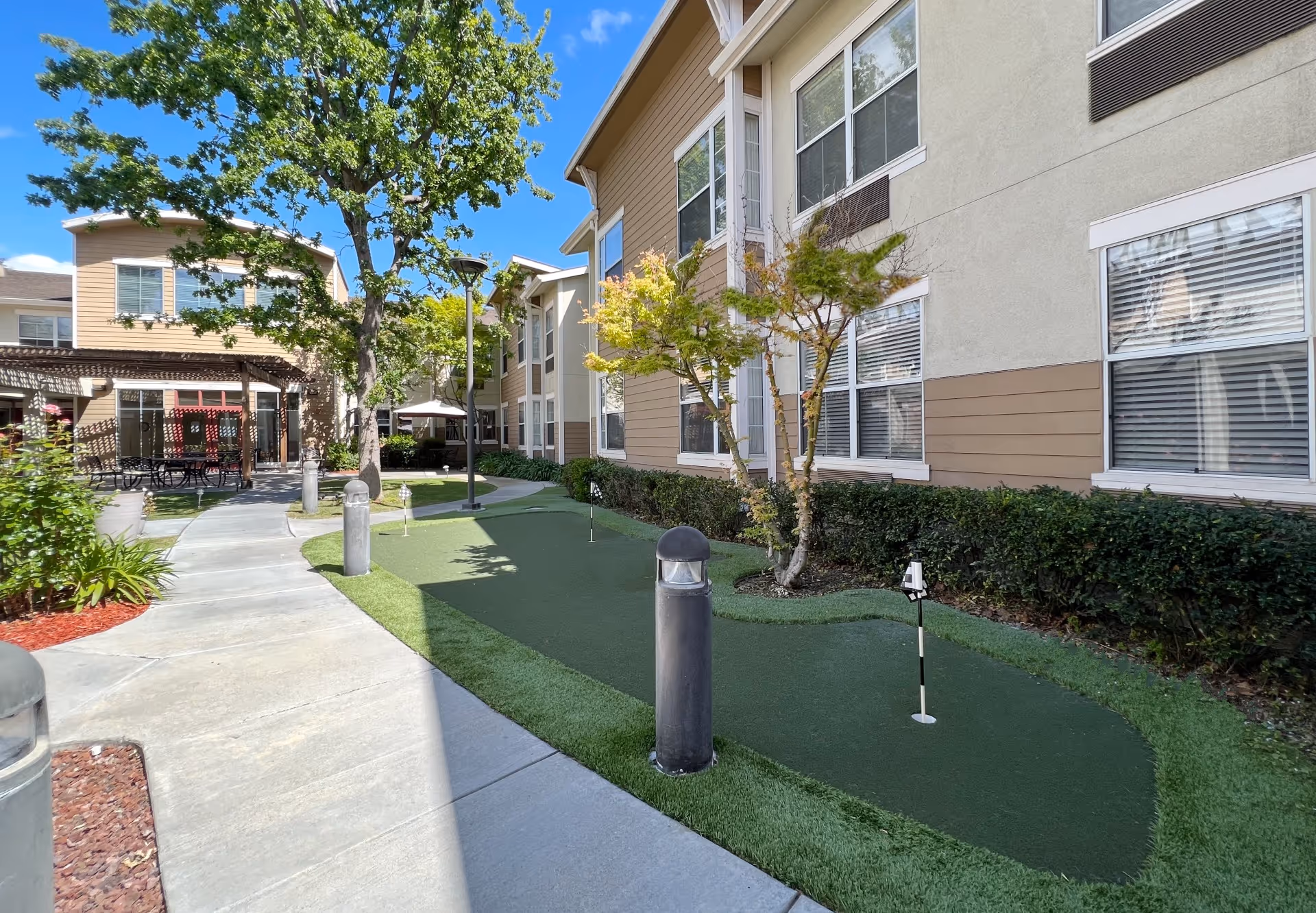 Outdoor courtyard area of a senior living facility with a putting green, trees, shrubs, and a paved walkway. The building has beige and tan siding with multiple windows. There are outdoor tables and chairs under a pergola in the background, and the sky is clear and blue.