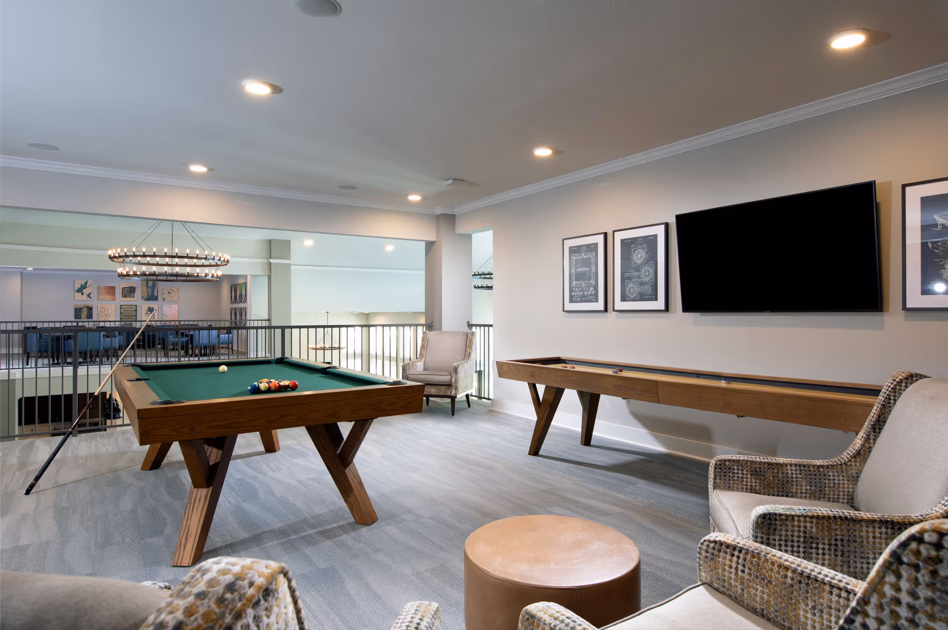 A bright game room with a pool table, shuffleboard, wall-mounted TV, and seating overlooking a lower level.