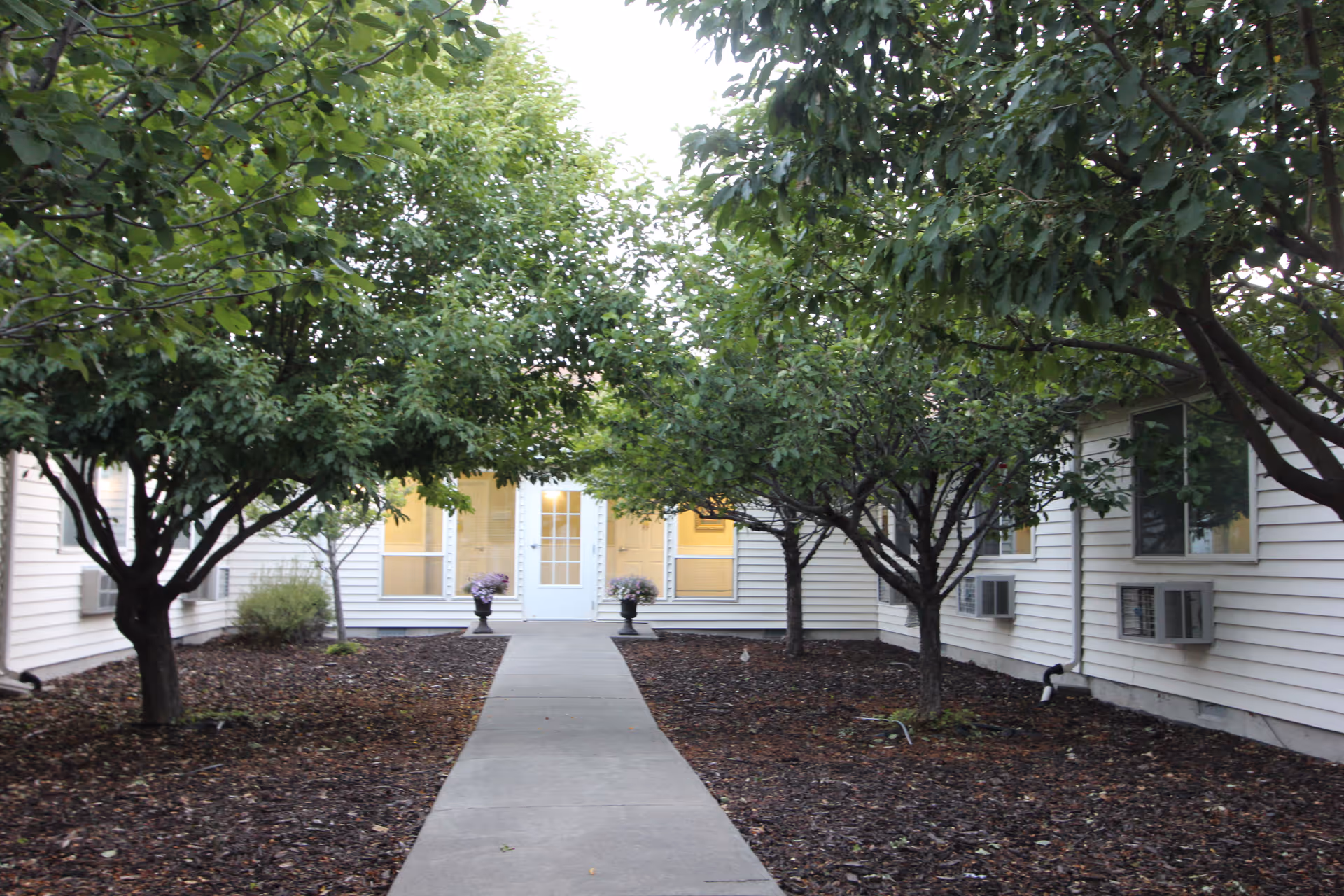 A paved walkway leading to a white door entrance of a building, flanked by two potted plants with flowers. The walkway is bordered by mulch beds with several leafy green trees on both sides. The building has white siding and multiple windows with air conditioning units installed.
