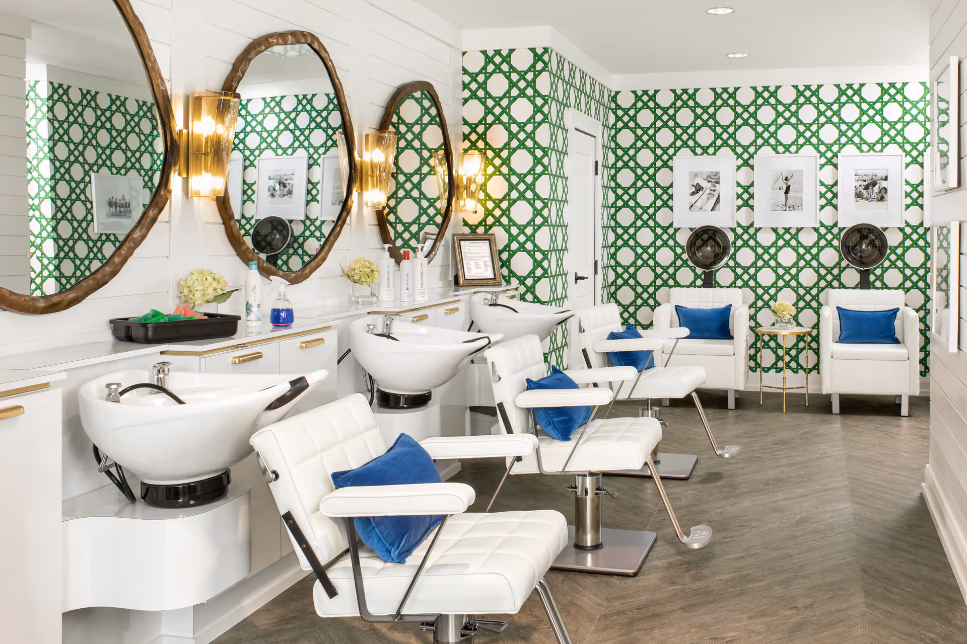 Interior of a stylish salon area with white salon chairs and wash basins. The walls feature a green and white patterned wallpaper, three large round mirrors with wooden frames, and wall-mounted lights. There are two white armchairs with blue cushions and a small round table between them, along with framed black and white photos on the wall.