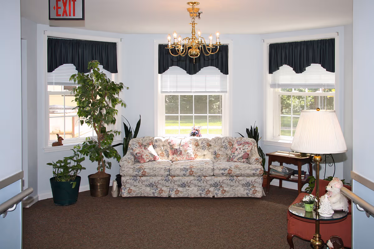 A cozy sitting area with a floral patterned sofa placed against three windows with white blinds and dark valances. There are several potted plants around the sofa, a small wooden side table with magazines, and a round glass-top table with a lamp and decorative items. A brass chandelier hangs from the ceiling, and an exit sign is visible above a doorway on the left.