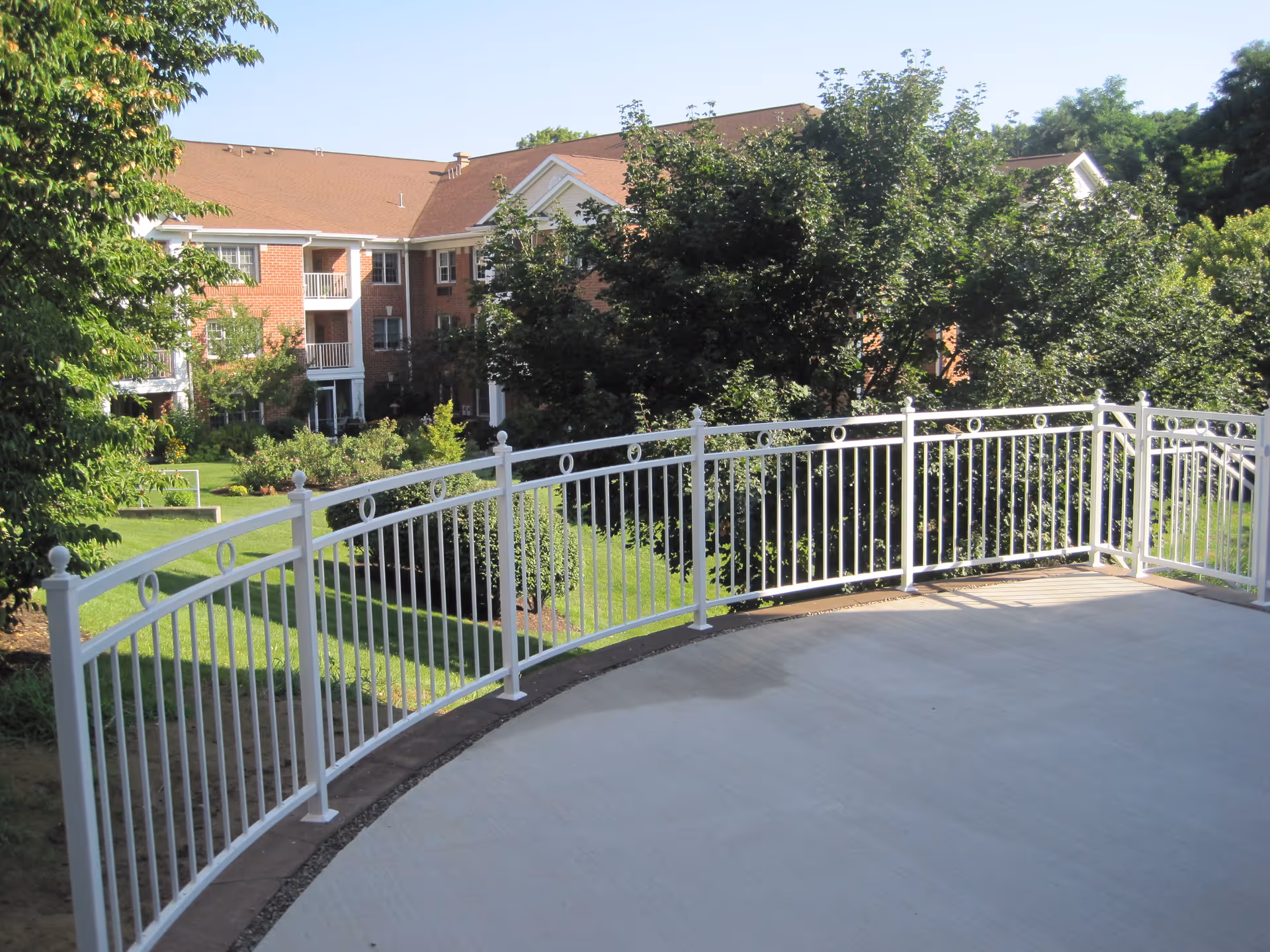 Outdoor patio area with a white metal railing overlooking a landscaped garden with green trees and bushes. In the background, there is a multi-story brick building with balconies and a red roof under a clear blue sky.