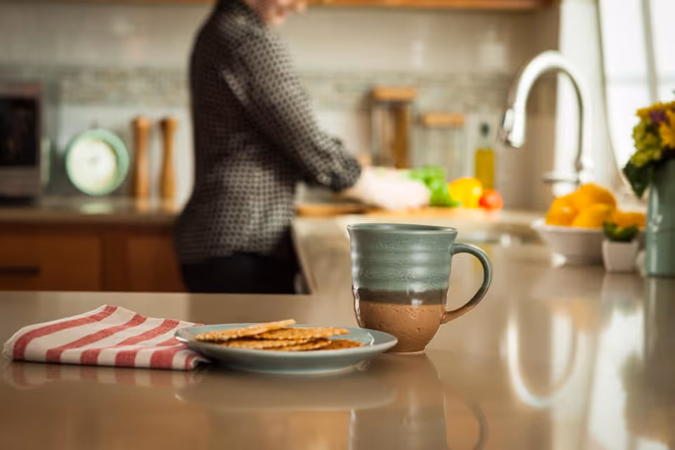 A ceramic mug and a plate with crackers on a kitchen counter with a red and white striped cloth. In the background, a person is preparing food near a sink with fresh vegetables and kitchen items visible.