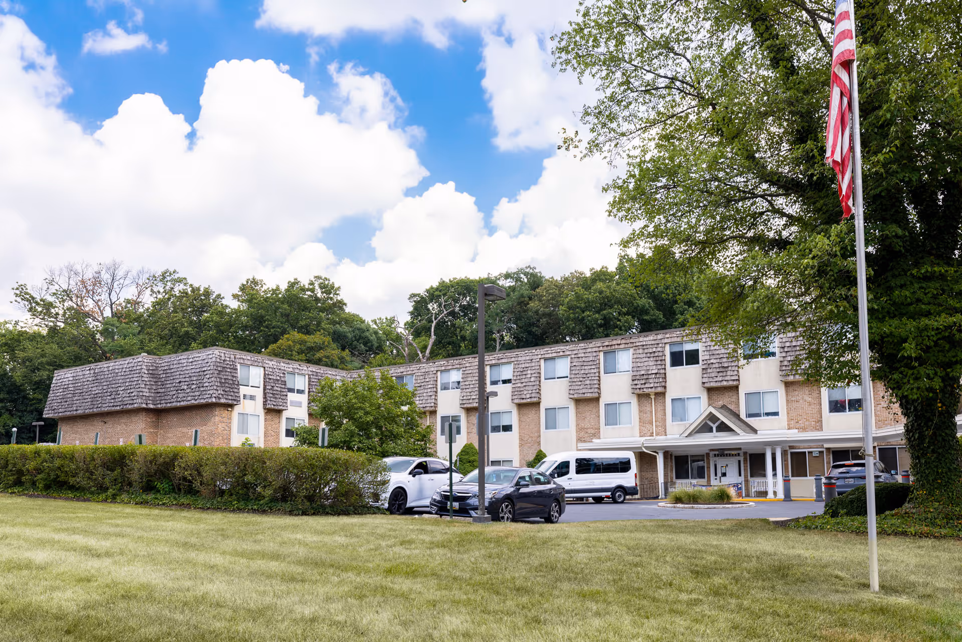 Exterior view of a multi-story senior living facility building with a shingled roof and beige brick walls. Several cars are parked in front of the entrance, and an American flag is visible on a flagpole to the right. The sky is partly cloudy with blue patches, and there are trees and bushes surrounding the building.