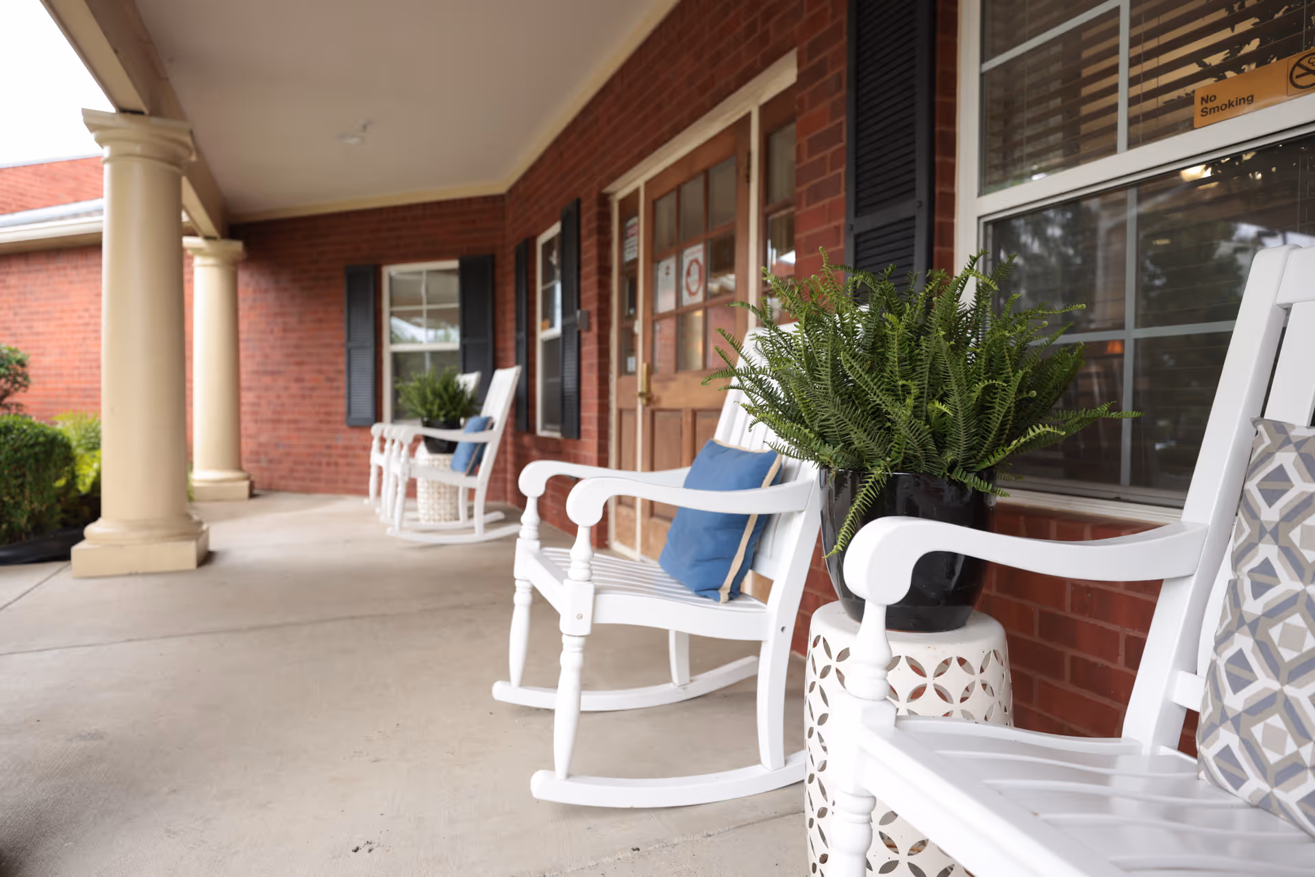Covered outdoor porch area with white rocking chairs adorned with blue and patterned cushions, potted green plants on small white tables, and a brick wall with windows and a wooden door in the background.