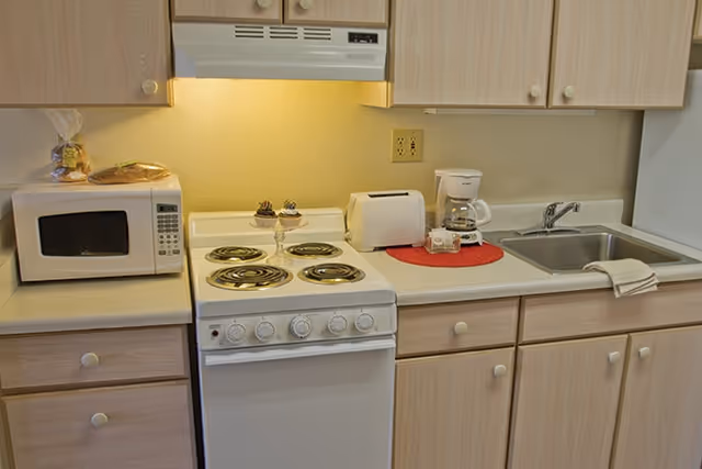 A compact kitchen area with light wood cabinets, a white electric stove with four coil burners, a microwave on the left countertop, a toaster and coffee maker on the right countertop, and a stainless steel sink with a white towel draped over the edge. There is a small decorative dish with succulents on the stove and a loaf of bread on top of the microwave.