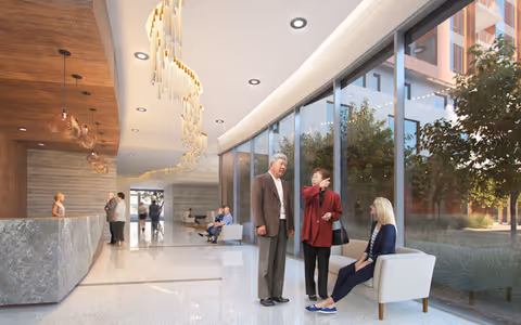 A modern senior living facility lobby with large floor-to-ceiling windows on the right side. Three seniors are interacting near the windows, with one woman sitting on a white chair and two others standing and talking. The lobby features a long reception desk on the left, a polished floor, contemporary lighting fixtures hanging from the ceiling, and additional seating areas further inside.