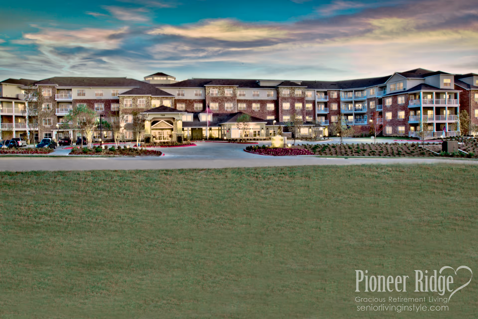 Wide exterior view of a large multi-story retirement living facility named Pioneer Ridge Gracious Retirement Living, with a spacious driveway, landscaped gardens, and balconies on the building. The sky is partly cloudy during sunset or dusk.