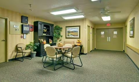 Carpeted common room with a round table and four chairs, a black cabinet with décor, potted plants, wall art, and double exit doors.