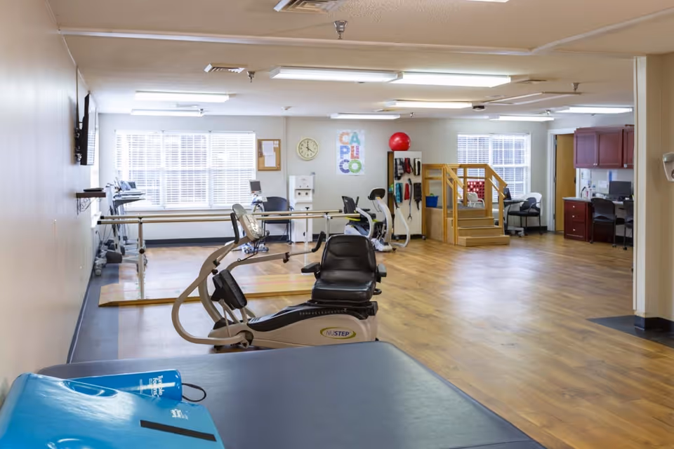 A spacious physical therapy room with exercise equipment including a recumbent bike, parallel bars, therapy stairs, and various exercise tools. The room has large windows letting in natural light, wooden flooring, and a small office area with desks and chairs in the back.