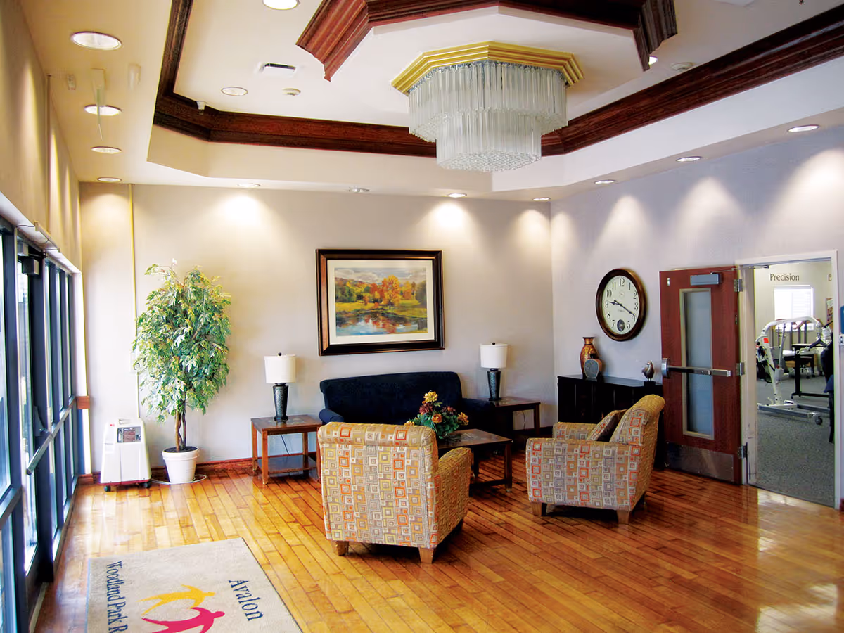 A well-lit lounge area with wooden flooring, two patterned armchairs, a dark blue sofa, two side tables with lamps, a coffee table with a flower arrangement, a large wall clock, a framed landscape painting, and a potted plant near glass doors. The ceiling features recessed lighting and a large chandelier. A door on the right leads to a room with exercise equipment.