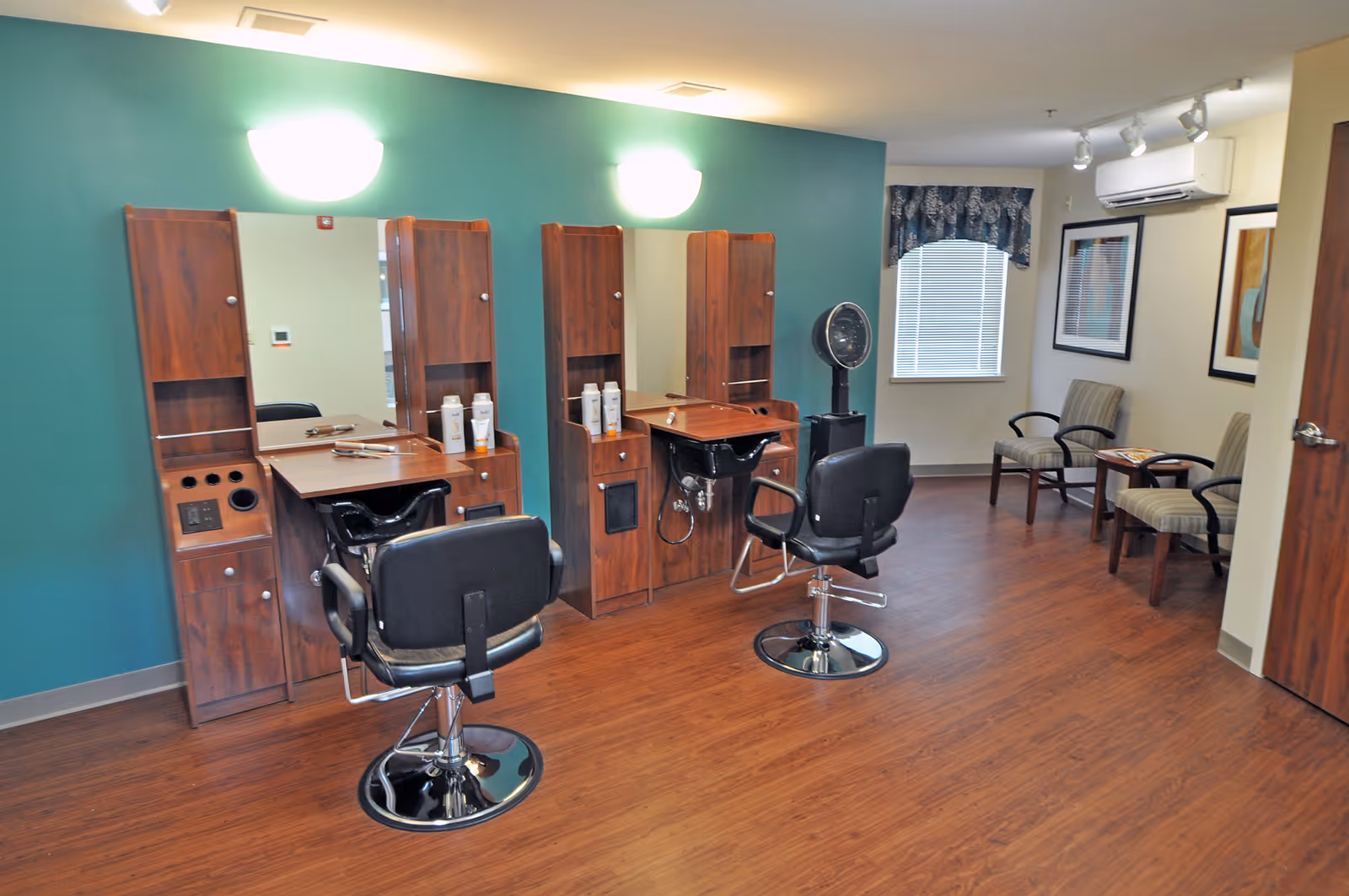 Interior view of a hair salon area in a senior living facility with two styling stations featuring mirrors, wooden cabinetry, and black salon chairs. There are two waiting chairs with a small table near a window with blinds and a valance. The floor is wooden, and the walls are painted teal and white.