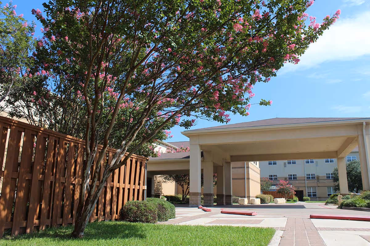 Entrance of a senior living building with a covered porte-cochere, a flowering crepe myrtle tree, wooden fence and landscaped lawn.