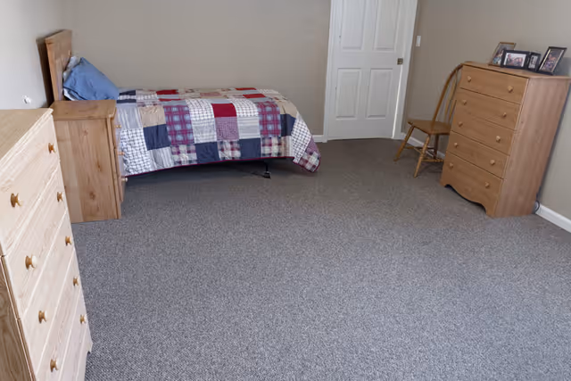 A simple bedroom with a single bed covered by a patchwork quilt in red, blue, and white colors. The room has beige walls and gray carpet flooring. There are two wooden dressers, one on the left and one on the right side of the room, with a wooden chair next to the dresser on the right. A few framed photos are placed on top of the right dresser. A closed white door is visible on the far wall.
