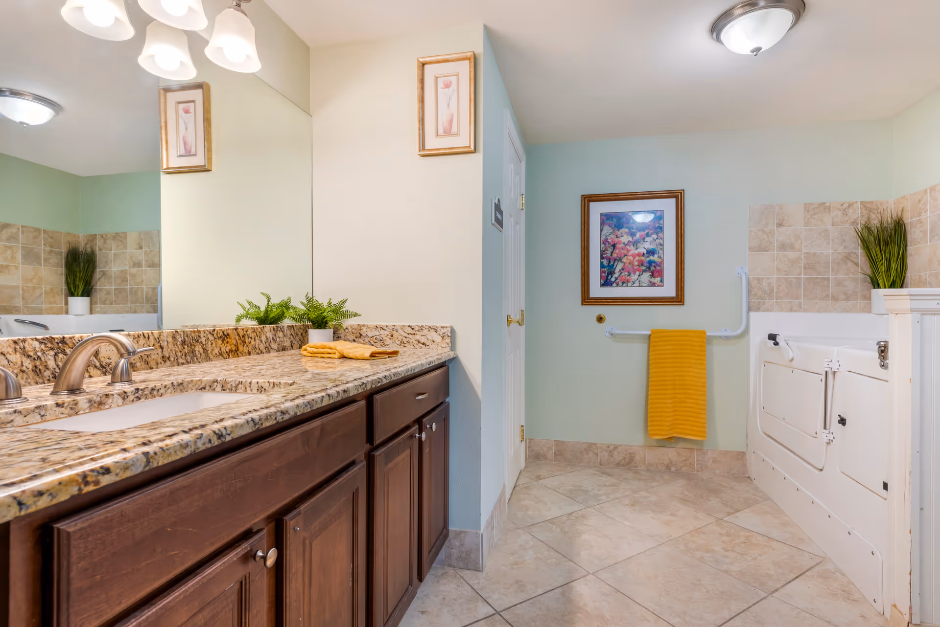 A bathroom with a granite countertop and dark wooden cabinets. There is a large mirror above the sink, a small potted plant, and folded yellow towels on the counter. The room features a walk-in bathtub with safety rails, beige tiled walls and floor, a framed floral picture on the wall, and a yellow towel hanging on a grab bar.