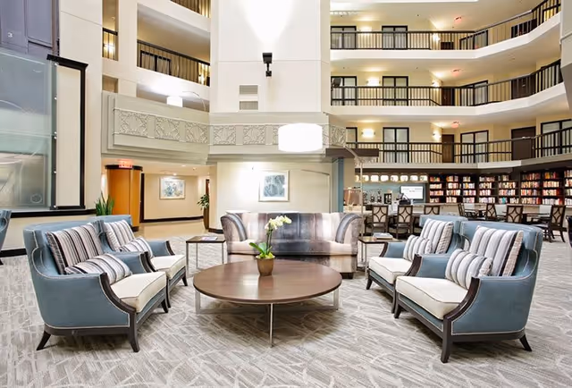 A spacious and well-lit common area in a senior living facility featuring a circular arrangement of comfortable upholstered chairs and sofas around a low round wooden coffee table with a small potted plant. The background shows multiple floors with railings overlooking the area, a bookshelf filled with books, and soft lighting fixtures.
