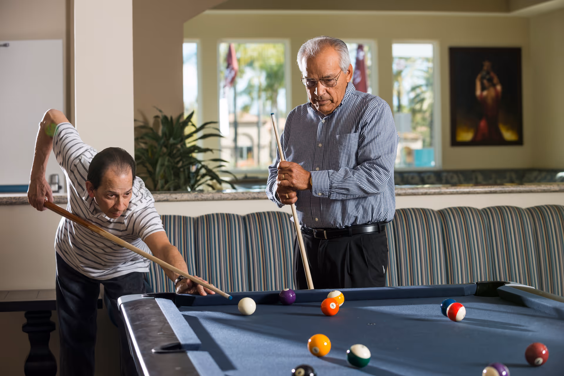 Two elderly men playing pool in a well-lit room with large windows and a striped cushioned bench in the background. One man is aiming a shot while the other watches, holding a pool cue.