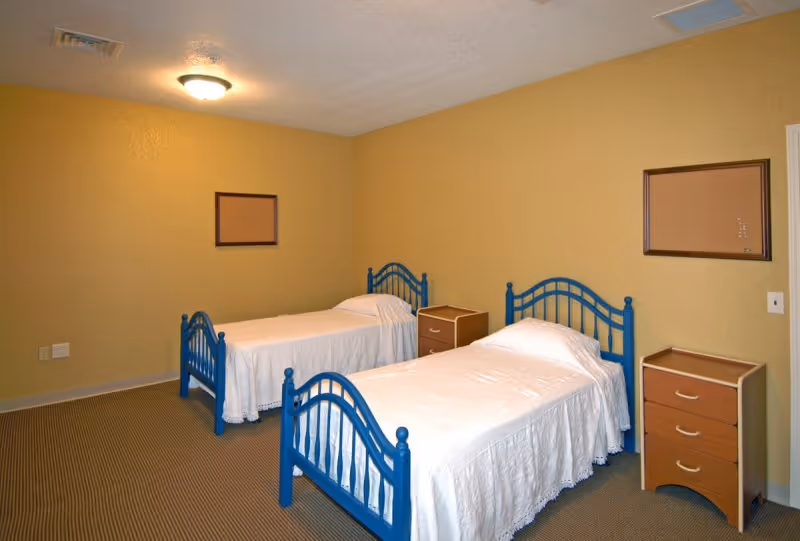 Simple bedroom with two twin beds with blue metal frames, white bedspreads, two bedside dressers, and corkboards on beige walls.