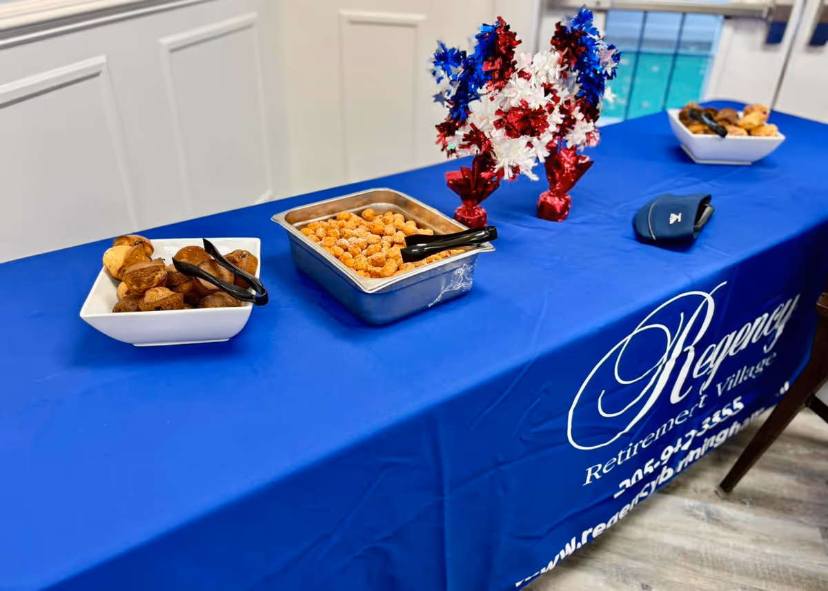 A table covered with a blue tablecloth featuring the Regency Retirement Village of Birmingham logo and contact information. On the table are two white bowls with muffins and a metal tray with tater tots, each with serving tongs. There are also red, white, and blue decorative pom-poms in the center of the table. The setting appears to be indoors with a window in the background.