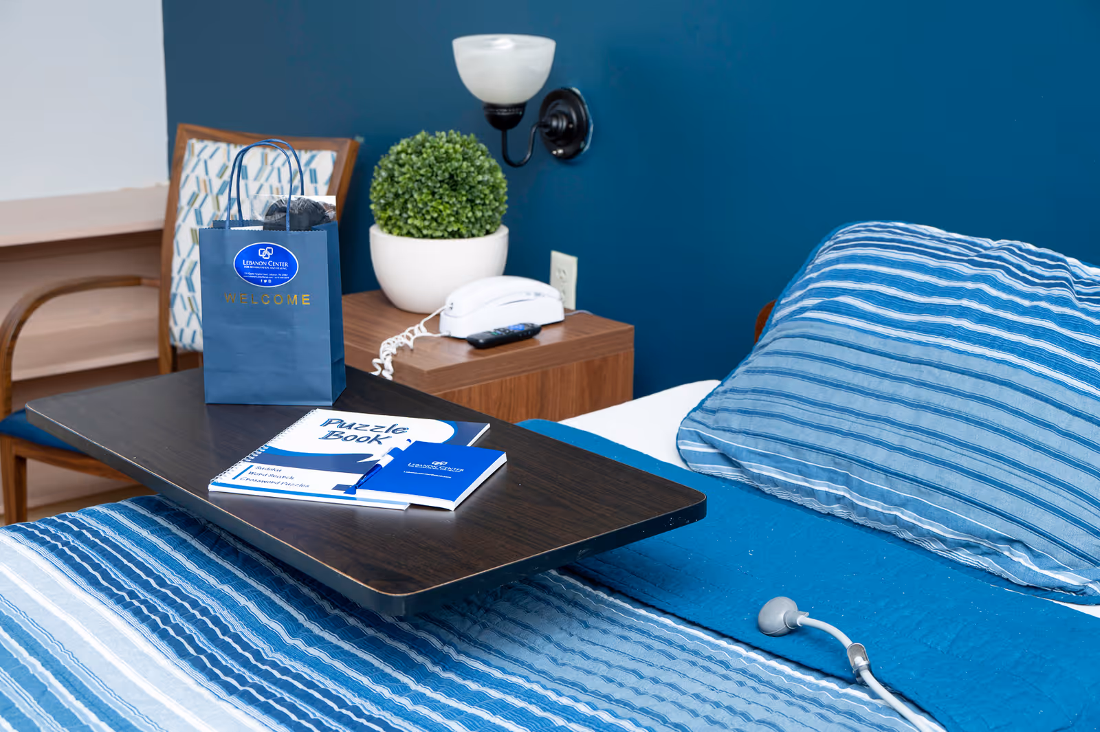 A neatly made single bed with blue striped bedding, an overbed table holding a welcome bag and puzzle book, and a bedside table with a phone and potted plant.