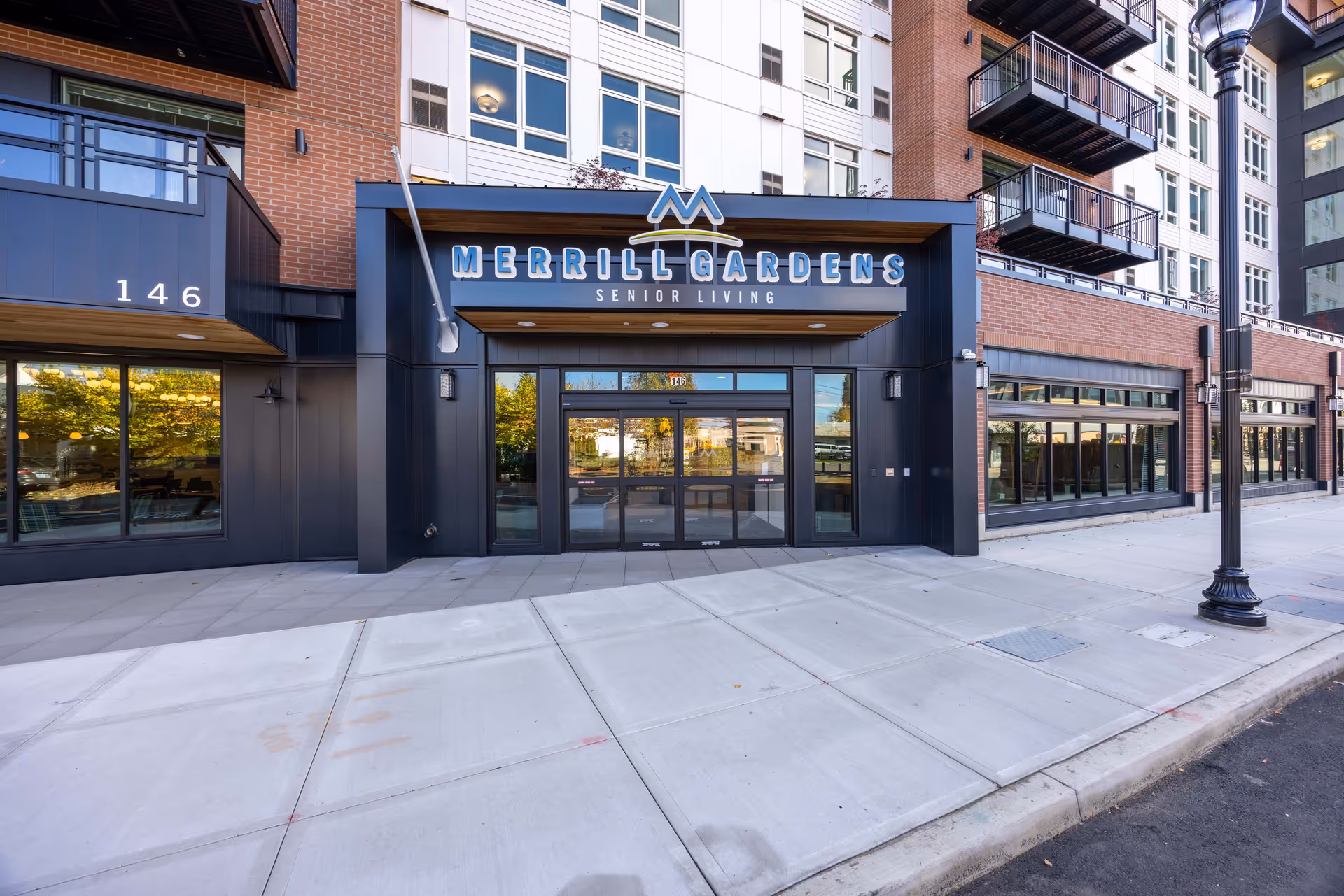 Exterior front entrance of Merrill Gardens senior living facility in Hillsboro, featuring a modern building facade with large windows, balconies, and a sidewalk in front.