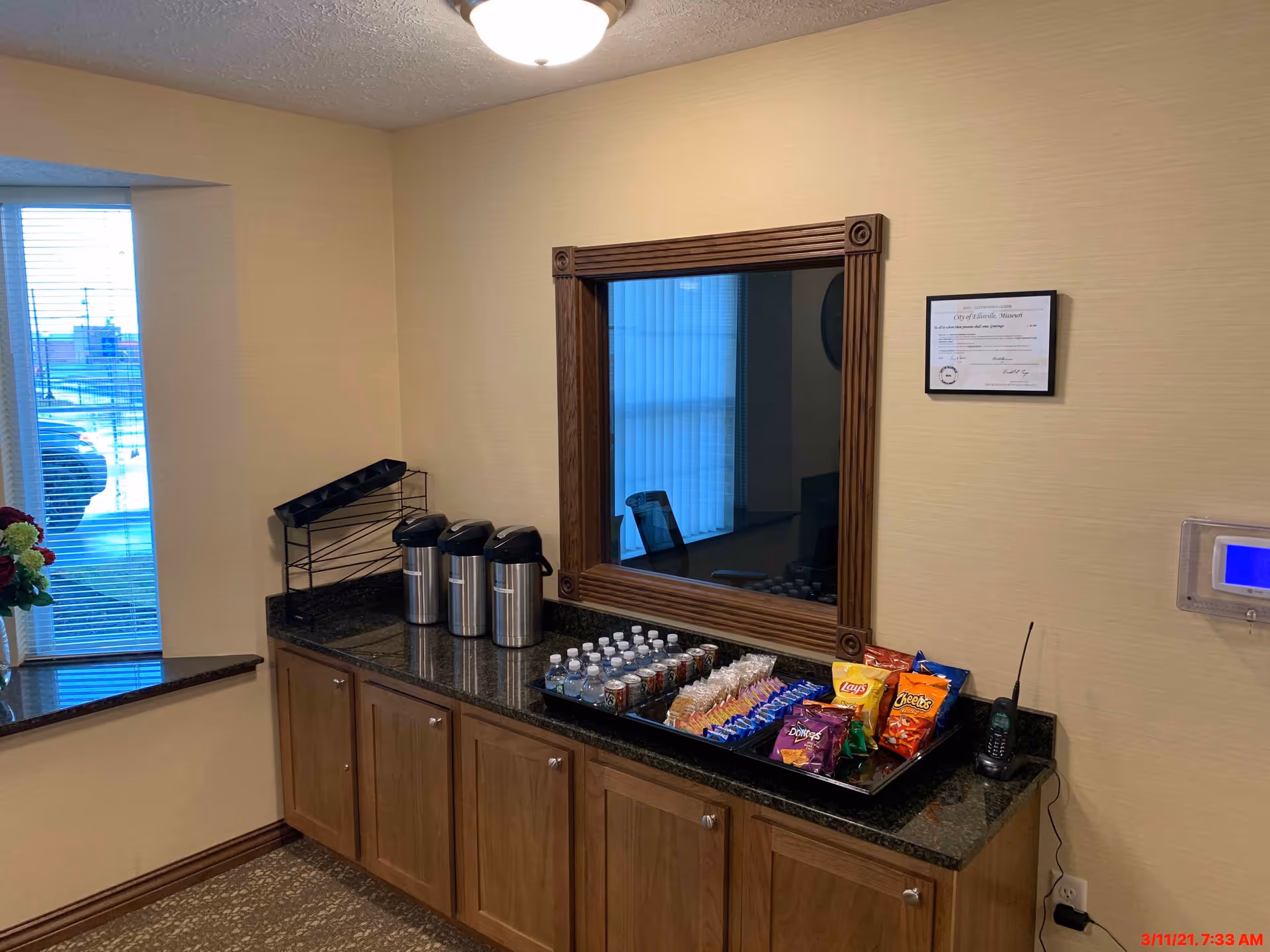 Snack and beverage station with coffee urns, bottled water, and packaged snacks on a granite countertop beneath a framed mirror in a communal area.