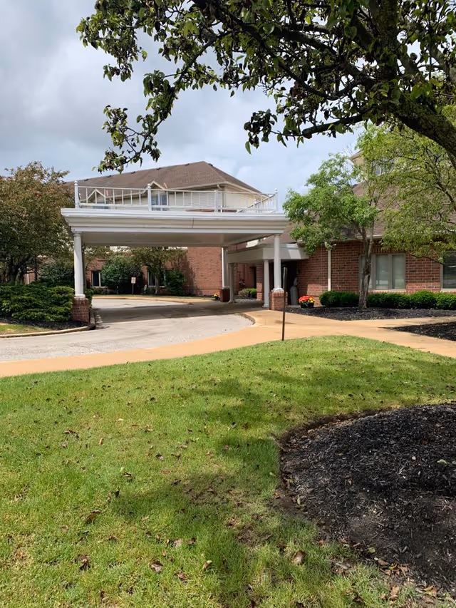 View of the exterior entrance area of a senior living facility with a covered drop-off area supported by white columns. The building is made of red brick with windows and surrounded by green grass, trees, and shrubs under a cloudy sky.