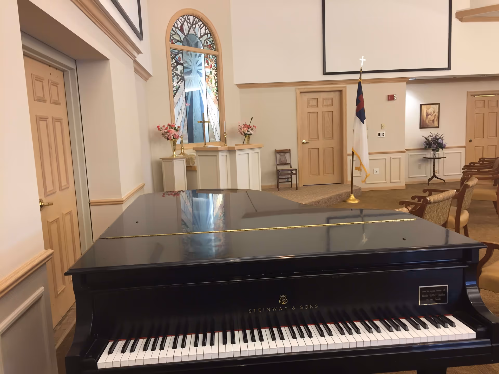 Interior view of a chapel or worship room with a black Steinway & Sons grand piano in the foreground. In the background, there is a white podium with a cross, two flower arrangements, a stained glass window with a tree design, a Christian flag on a stand, wooden chairs, and beige walls with wooden doors.