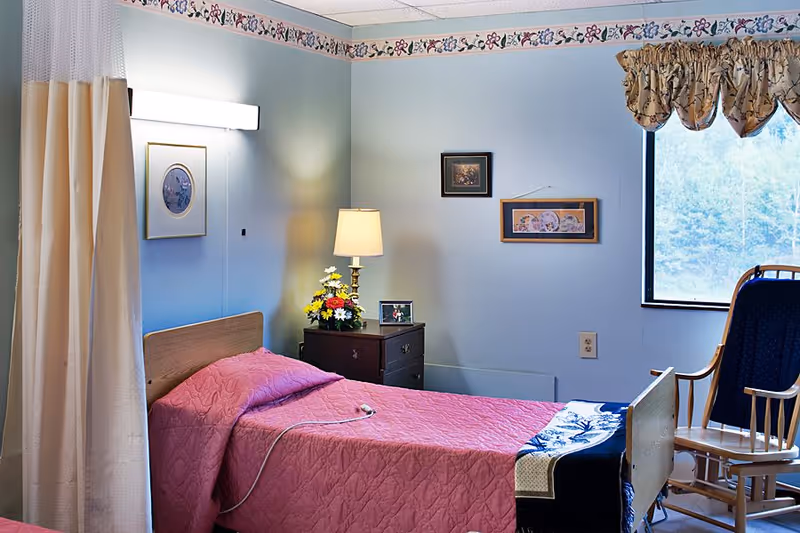 Small, softly lit resident bedroom with a single bed covered by a pink quilt, a nightstand with a lamp and flowers, wall art, a window, and a rocking chair.