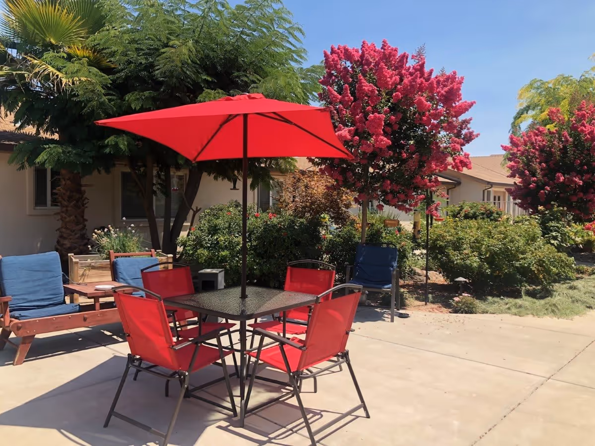 Outdoor patio area with a square glass table surrounded by four red chairs and a large red umbrella providing shade. There are additional wooden chairs with blue cushions nearby. The patio is surrounded by green bushes and trees, including vibrant pink flowering trees, under a clear blue sky.