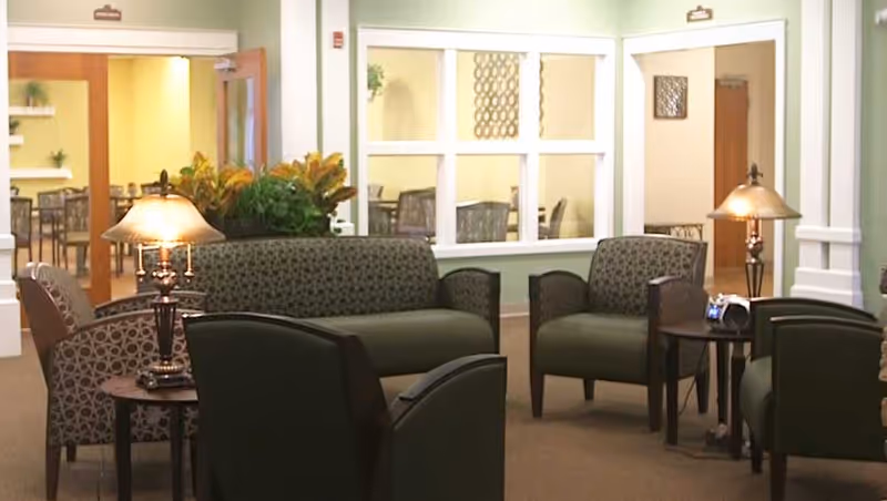 A cozy senior living facility common area with patterned armchairs and a loveseat arranged around small wooden tables with lamps. There are plants and a window looking into another room with more chairs and tables.