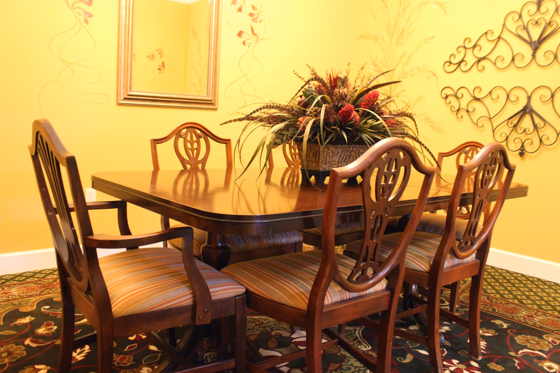 A dining room with a wooden dining table surrounded by six wooden chairs with striped cushions. A decorative floral arrangement is placed in the center of the table. The walls are painted yellow and decorated with a framed mirror and ornamental metal wall art. The floor is covered with a patterned carpet.